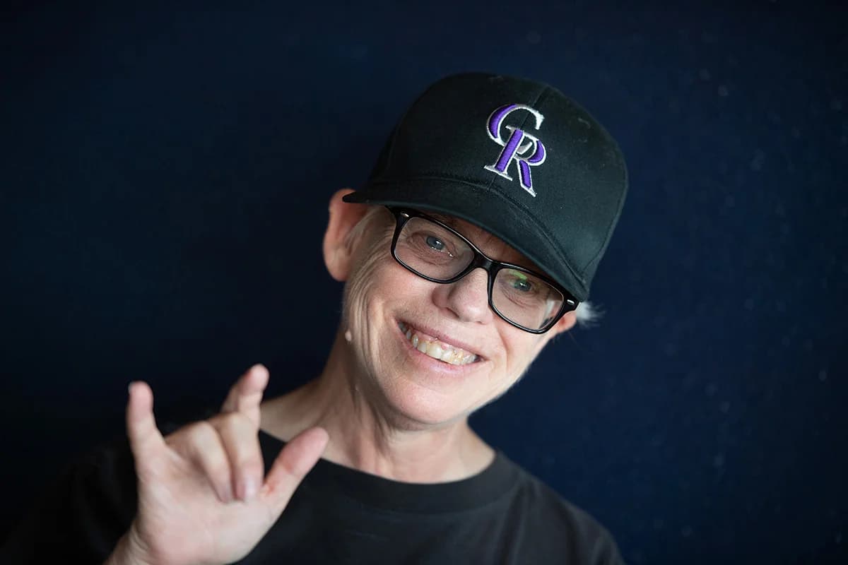 A smiling woman in glasses and a Colorado Rockies cap makes the “I love you” sign.