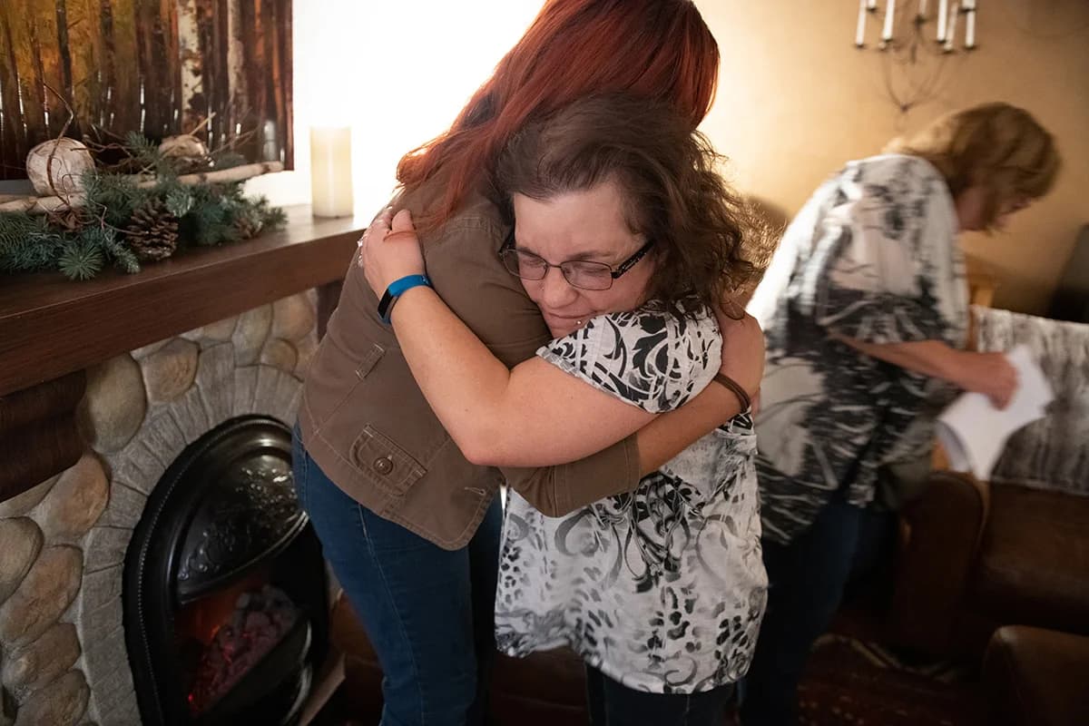 Two women share a warm hug in a cozy room beside a stone fireplace.