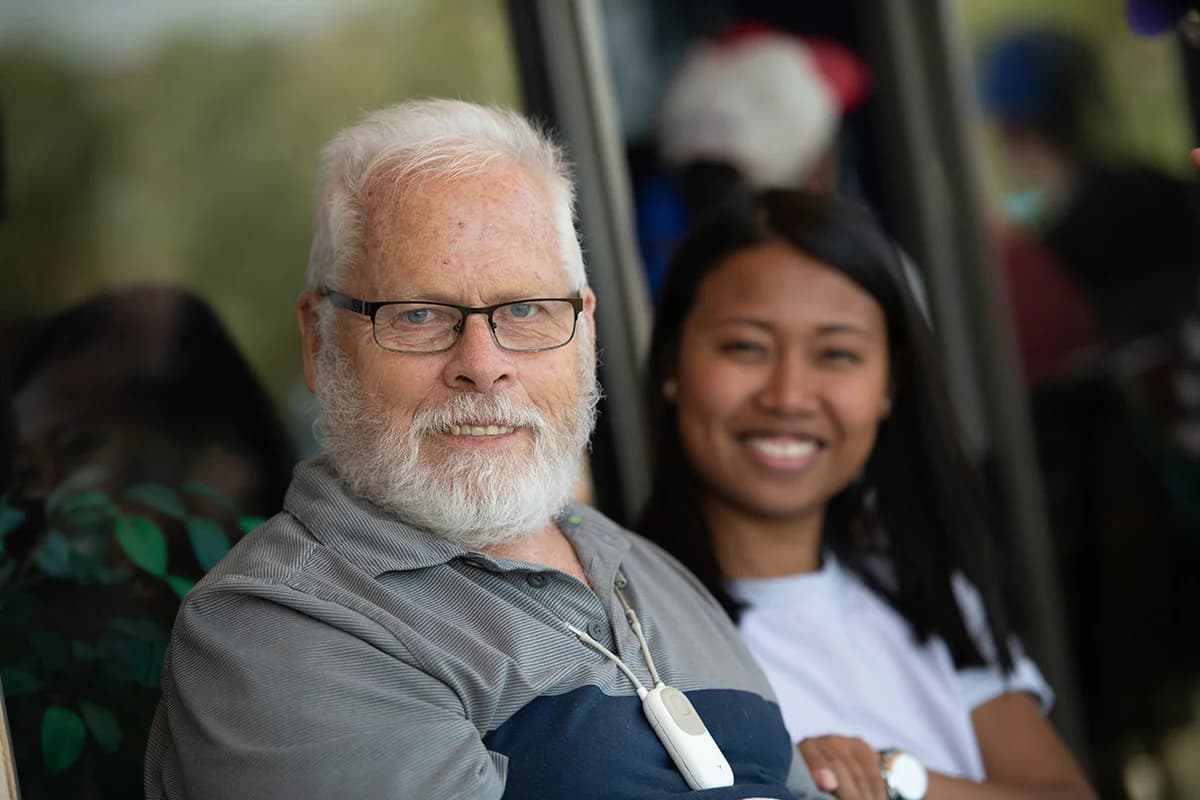 An older man with a white beard sits beside a smiling young woman outdoors.